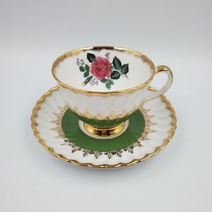 May include: A vintage teacup and saucer set. The white teacup features a pink rose design and gold trim. The saucer has a green center and white scalloped edges with gold accents. A classic tea set.