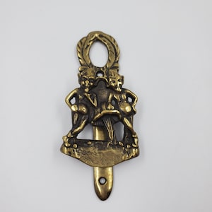 Peut inclure: Un heurtoir de porte en laiton avec un motif de trois personnages se tenant la main. Le heurtoir a un anneau en haut et un crochet en bas.