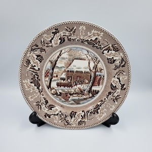 May include: A brown and white ceramic plate featuring a winter scene with a cabin, trees, and a horse-drawn wagon. The plate has a decorative border of oak leaves and acorns.