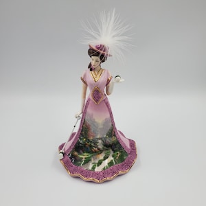 May include: A porcelain figurine of a woman in a pink Victorian-style dress and hat, with white feather detailing. The dress features a landscape painting. The figurine is holding a small object and a white parasol.
