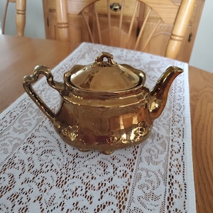 May include: A gold teapot with a lid and a handle. The teapot is sitting on a white lace tablecloth.