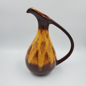 May include: A vintage ceramic jug with a teardrop shape. The jug features a dark brown handle and spout, with a gradient of yellow and brown tones on the body. The base is a solid dark brown colour.
