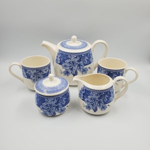 May include: A ceramic tea set comprising a teapot, a lidded sugar bowl, a creamer, and two mugs. Each piece is off-white with a blue transferware pattern of grape leaves and clusters, finished with a decorative chain-link border around the rims.