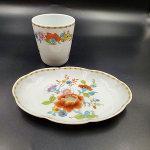 May include: A white porcelain teacup and saucer set with a floral design. The cup has a scalloped rim and the saucer has a wavy edge. The flowers are in shades of orange, pink, blue, and green.