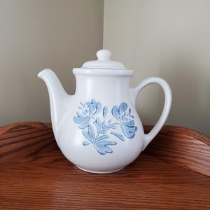 May include: White ceramic teapot with a blue floral design. The teapot has a round body and a spout on one side and a handle on the other.