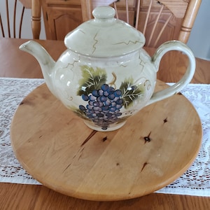 May include: A ceramic teapot with a hand-painted design of grapes and leaves. The teapot is a light beige colour with a glossy finish. It is sitting on a wooden base.