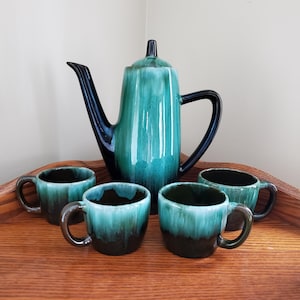 May include: A vintage ceramic teapot and four matching cups in a teal and black glaze. The teapot has a tall, slender body and a spout on the side. The cups are small and round with a handle on the side.