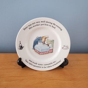 May include: A white ceramic plate featuring a scene with a rabbit in bed, with text around the rim. The plate has a teapot and teacup illustration. The text reads: "Peter was not very well during the evening. His mother put him to bed, and made some camomile tea: One table-spoonful to be taken at bed-time."