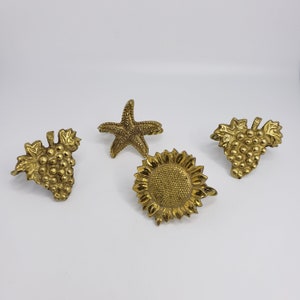 Peut inclure: Quatre anneaux de serviette en métal doré avec des motifs différents : une étoile de mer, un tournesol et deux grappes de raisin.