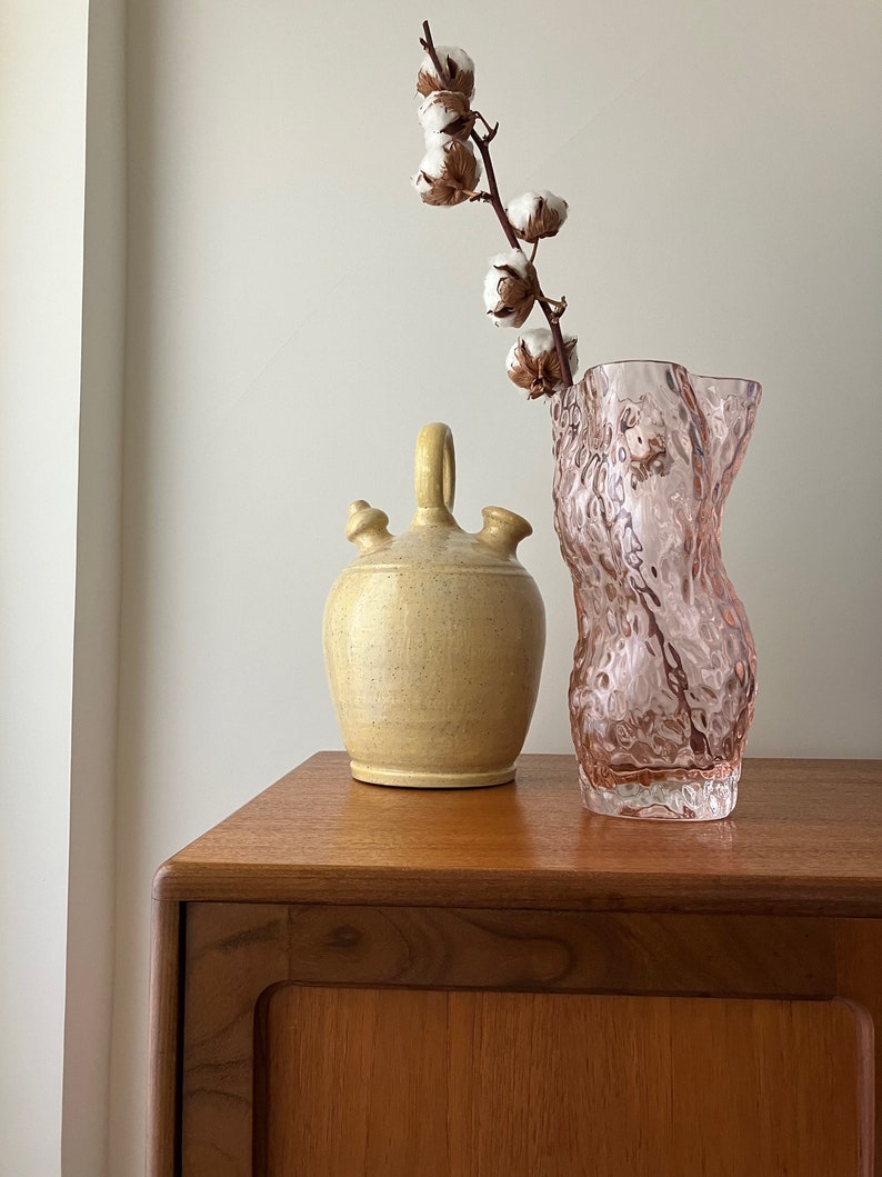 May include: A light pink glass vase with a textured surface, a yellow ceramic pitcher with a spout, and a branch of white cotton flowers on a wooden table.