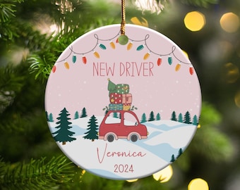 May include: A round ceramic Christmas ornament featuring a red car carrying a stack of gifts on its roof, set against a snowy landscape with pine trees. The text reads NEW DRIVER, Veronica, and 2024, with a string of colorful holiday lights above.