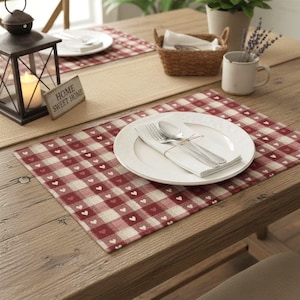May include: A rectangular placemat featuring a red and white checkered pattern with small white hearts inside the squares. A white ceramic plate sits on the placemat, topped with a white folded napkin, a silver fork, and a silver spoon.