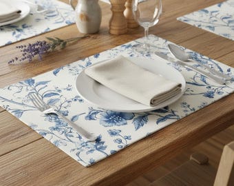 May include: A rectangular placemat featuring a blue and white floral pattern, topped with a white ceramic plate, a folded beige cloth napkin, a silver fork, a silver knife, and a silver spoon, arranged on a wooden table surface.
