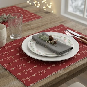May include: A rectangular red placemat featuring a repeating white script pattern of the word joy and small hearts. The setting includes a white plate, a gray napkin topped with a small pinecone and greenery, a white mug, and a glass.
