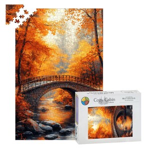 May include: A jigsaw puzzle featuring an autumn forest scene with a stone bridge over a stream. The puzzle box is white with the text Cozy Cabin Gifts Co and The World's Most Beautiful Jigsaw Puzzles printed on the front and top edges.