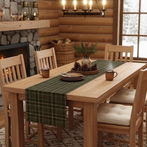 May include: A wooden dining table set with four chairs featuring cream-colored cushions. The table is decorated with a dark green plaid table runner, two brown ceramic mugs, a stack of plates, and a centerpiece of pinecones in a round tray.