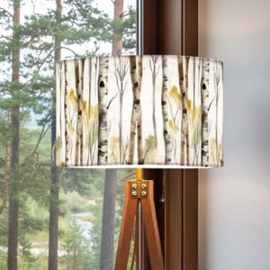 May include: A floor lamp featuring a cylindrical drum shade decorated with a watercolor-style print of birch trees in shades of white, brown, and yellow. The lamp base is constructed from three wooden legs joined by metal hardware.