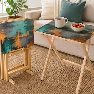 May include: Set of four folding TV trays with a colorful abstract design in teal, orange, and gray. The trays have a natural wood frame and are set up in a living room setting. One tray is set up with a cup of coffee and a bowl of strawberries.