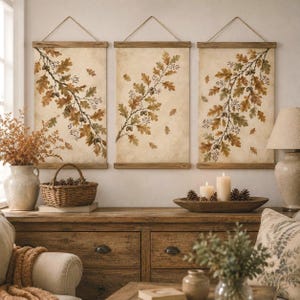May include: Three vertical wall hangings featuring botanical illustrations of oak branches with brown and green leaves and acorns. Each print is mounted on wooden rails with a twine hanger, displaying a neutral background with scattered leaves.