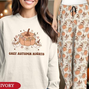 May include: An ivory long-sleeve sweatshirt and matching pyjama trousers featuring a repeating pattern of orange pumpkins adorned with small flowers, stars, and crescent moons. The sweatshirt displays the text COZY AUTUMN NIGHTS in brown.