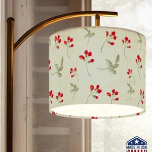 May include: A floor lamp with a curved, gold-toned metal frame. The lampshade is cylindrical, featuring a cream-colored background with a repeating pattern of red berries and green leaves. The lamp is marked "Made in USA".