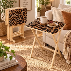 May include: A set of four folding snack tables with light-colored wooden frames and rectangular tops featuring a brown and black leopard print pattern. One table is unfolded, holding a white ceramic mug and a small plate with cookies.