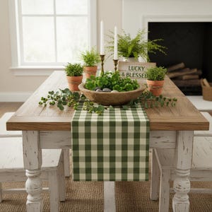 May include: A wooden dining table with a green and white checkered table runner. The table features a wooden bowl filled with green clover, two white taper candles in metal holders, three potted plants, and a sign that reads LUCKY & BLESSED.