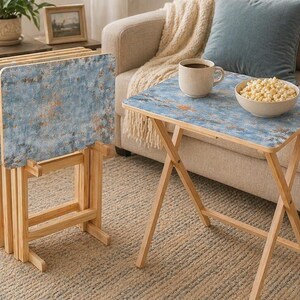 May include: A set of four folding snack tables with light wood frames and rectangular tops featuring a distressed blue and copper-toned finish. One table is set up for use, while the other three are folded and stored in a matching wooden stand.