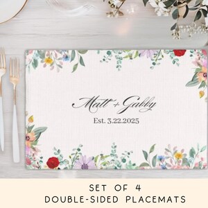 May include: A rectangular placemat featuring a white background with a colorful floral border of red, yellow, purple, and green foliage. The center displays the text Matt + Gabby and Est. 3.22.2025 in black script and serif fonts.