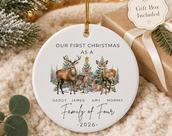 May include: A round white ceramic Christmas ornament featuring a printed illustration of four deer in a snowy forest. The text reads: OUR FIRST CHRISTMAS AS A, DADDY JAMES AMY MOMMY, Family of Four, and 2026. A gold string is attached to the top.