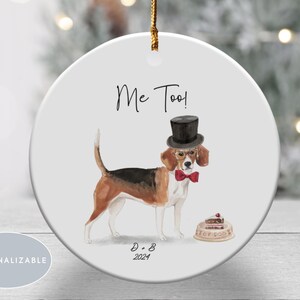 May include: A round white ceramic ornament featuring a watercolor illustration of a beagle wearing a black top hat, glasses, and a red bow tie. The text Me Too! is above the dog, with D + B 2024 below. A small cake sits on a plate nearby.