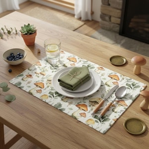 May include: A rectangular placemat featuring a watercolor-style print of various mushrooms and green foliage. Centered on the placemat is a white ceramic plate topped with a folded olive green cloth napkin tied with twine, a silver knife, and a spoon.