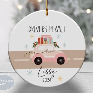 May include: A round white ceramic ornament featuring a pink car carrying colorful gift boxes on its roof. The text DRIVERS PERMIT is printed above the car, with the name Lissy and the year 2024 below. A string of lights is draped over the car.