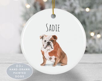 Puede incluir: Un adorno redondo de cerámica blanca con una ilustración en acuarela de un bulldog marrón y blanco. El nombre SADIE está impreso en texto negro sobre el perro. Un cordón dorado está sujeto en la parte superior. El adorno tiene un acabado liso.