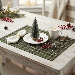 May include: A green and white plaid placemat featuring a white plate topped with a small artificial pine tree, a white napkin with a gold ring, a white mug filled with marshmallows, and decorative red berries and pinecones scattered around.