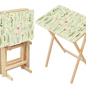 May include: A set of folding snack tables with light wood frames and rectangular tops featuring a pale green background with a botanical print of various wildflowers, grasses, and small pink and yellow blossoms in a repeating pattern.