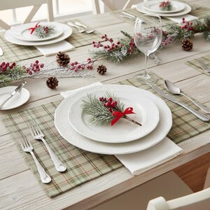 May include: A table setting featuring white ceramic plates, silver forks, a knife, and a spoon on a green and tan plaid placemat. A sprig of frosted pine needles with red berries and a red ribbon sits on the center plate. A clear glass goblet is present.