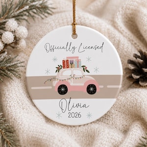 May include: A round white ceramic ornament hanging from a gold string. It features a pink car carrying colorful gift boxes on its roof, decorated with string lights. The text reads Officially Licensed, Olivia, and 2026, surrounded by snowflakes.