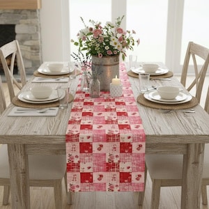 May include: A rectangular table runner featuring a patchwork design of alternating red and pink squares with floral and plaid patterns. The runner is centered on a wooden dining table set with white plates, clear glasses, and a floral centerpiece.