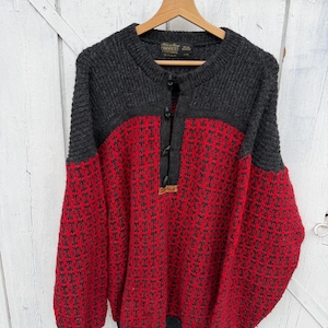 May include: A long-sleeve knit sweater featuring a charcoal gray upper section and a vibrant red lower section with a repeating black geometric pattern. The sweater has a round neckline with a partial button-down placket and a small brown brand tag.
