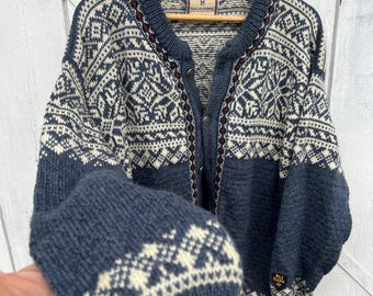 Può includere: Un cardigan blu e crema con un motivo geometrico. Il maglione ha una chiusura con bottoni sul davanti, uno scollo tondo e maniche lunghe. I polsini e l'orlo sono blu tinta unita e il maglione è appeso a una gruccia di legno.