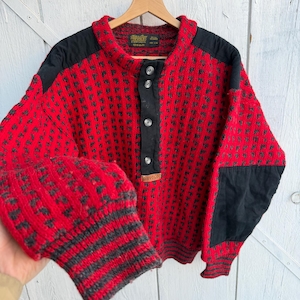 Puede incluir: Un suéter rojo y negro estampado con cuello alto y cierre de botones. El suéter tiene parches negros en los hombros y codos. Los puños tienen rayas grises y negras.