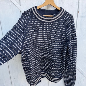 Puede incluir: Jersey azul marino con un patrón geométrico en blanco roto. El jersey tiene cuello redondo y puños y dobladillo acanalados. El diseño presenta un patrón repetitivo de pequeños cuadrados y rayas alrededor del cuello y el dobladillo.