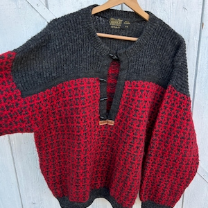 Puede incluir: Un suéter de lana gris y rojo con un diseño estampado. El suéter tiene cuello redondo y cierre de botones. Las mangas son largas y los puños son grises. El suéter está colgado en una percha de madera.