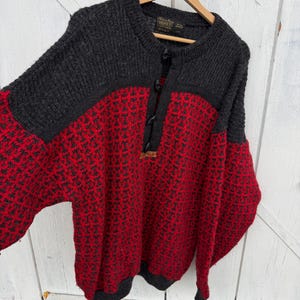 May include: A long-sleeve knit sweater featuring a charcoal gray upper section and a vibrant red and charcoal gray patterned lower body. The sweater has a round neckline with a partial button-down placket, black buttons, and a small Devold brand tag.