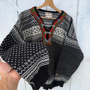 Könnte beinhalten: Ein schwarz-weißer Wollpullover mit komplexem geometrischem Muster, einem V-Ausschnitt mit roter, grüner und blauer Stickerei sowie zwei silbernen Metallverschlüssen. Der Pullover hat lange Ärmel mit Rippbündchen und ein Etikett von Dale of Norway.