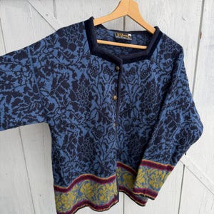 Peut inclure: Un cardigan bleu avec un motif floral et une encolure carrée. Le pull a une bordure bleu foncé et des boutons dorés. Les poignets et l'ourlet présentent une bande décorative avec des accents jaunes, rouges et verts. Le cardigan est suspendu à un cintre en bois.