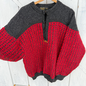 Puede incluir: Un suéter de lana vintage en rojo y gris, con un patrón texturizado y un cuello abotonado. La parte superior y las mangas son grises, mientras que el cuerpo es rojo con un diseño gris oscuro. El suéter cuelga de una percha de madera.