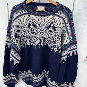 May include: A navy blue sweater with a cream-coloured geometric pattern across the chest and shoulders. The sweater has a crew neck and long sleeves. The cuffs, neckline, and hem feature a patterned border with red, green, and blue accents. The label reads "Dale of Norway".