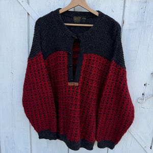 May include: A long-sleeve knit sweater featuring a dark charcoal gray upper section and a vibrant red and charcoal gray patterned lower section. The sweater has a partial front placket with three toggle closures and a small leather Devold brand tag.